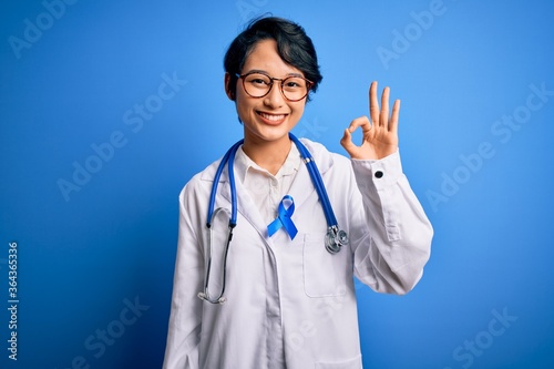 Young beautiful asian doctor girl wearing stethoscope and coat with blue cancer ribbon smiling positive doing ok sign with hand and fingers. Successful expression.