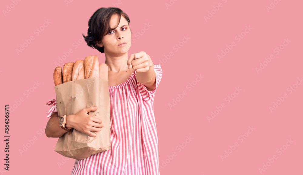 Beautiful young woman with short hair holding paper bag with bread pointing with finger to the camera and to you, confident gesture looking serious Beautiful young woman with short hair holding paper bag with bread pointing with finger to the camera and to you, confident gesture looking serious