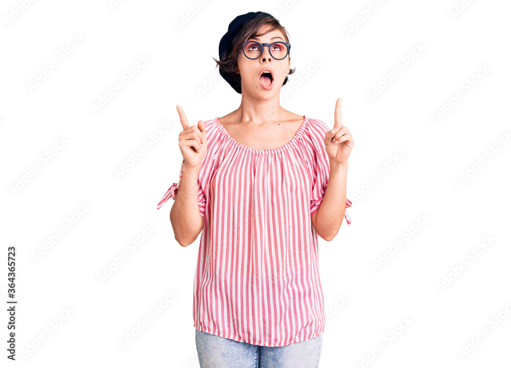 Beautiful young woman with short hair wearing casual clothes and glasses amazed and surprised looking up and pointing with fingers and raised arms. Beautiful young woman with short hair wearing casual clothes and glasses amazed and surprised looking up and pointing with fingers and raised arms.
