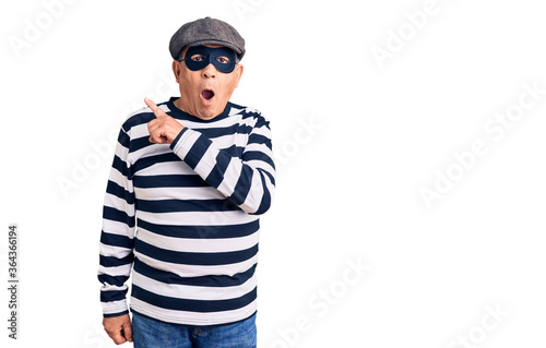 Senior handsome man wearing burglar mask and t-shirt surprised pointing with finger to the side, open mouth amazed expression.