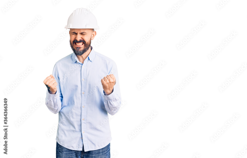 Young handsome man wearing architect hardhat very happy and excited doing winner gesture with arms raised, smiling and screaming for success. celebration concept. Young handsome man wearing architect hardhat very happy and excited doing winner gesture with arms raised, smiling and screaming for success. celebration concept.