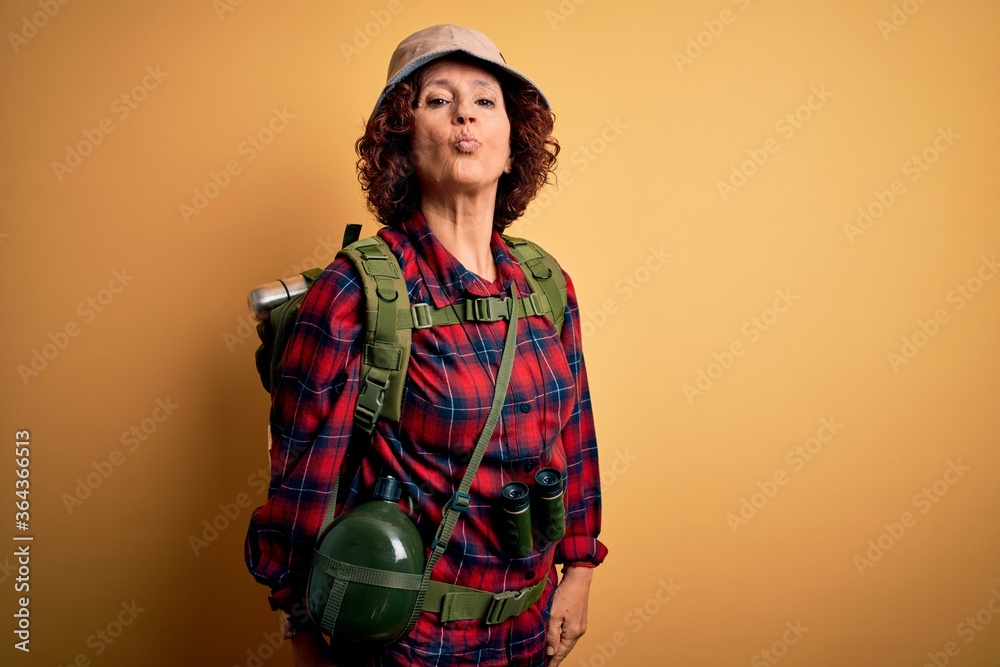 Middle age curly hair hiker woman hiking wearing backpack and water canteen using binoculars looking at the camera blowing a kiss on air being lovely and sexy. Love expression.