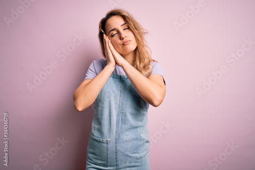 Young beautiful blonde woman wearing casual denim overalls standing over pink background sleeping tired dreaming and posing with hands together while smiling with closed eyes.