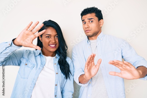 Beautiful latin young couple wearing casual clothes afraid and terrified with fear expression stop gesture with hands, shouting in shock. panic concept.