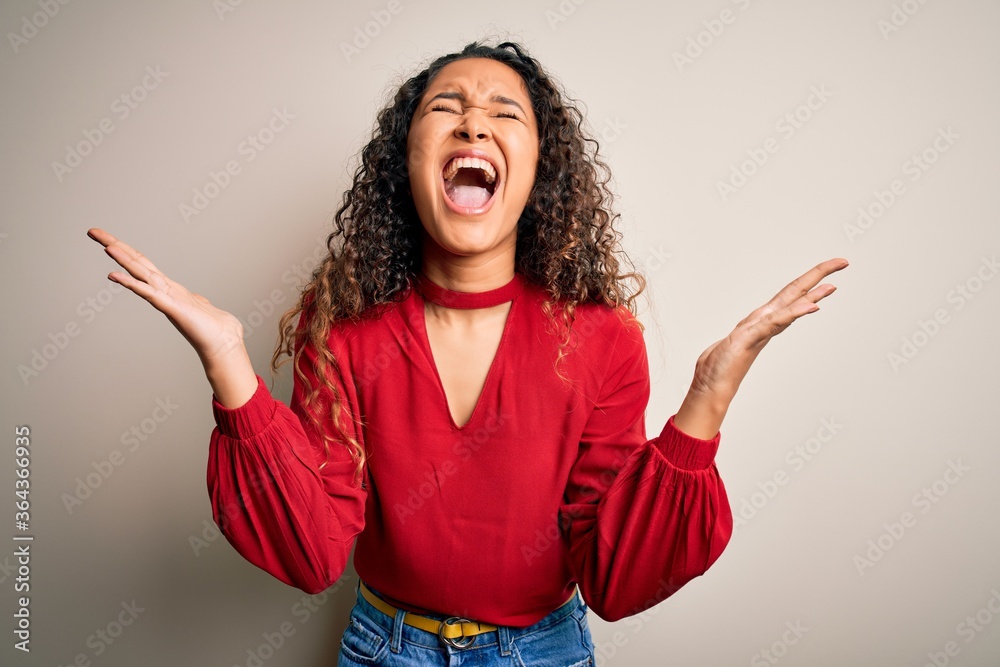 Young beautiful woman with curly hair wearing casual t-shirt standing over white background celebrating mad and crazy for success with arms raised and closed eyes screaming excited. Winner concept Young beautiful woman with curly hair wearing casual t-shirt standing over white background celebrating mad and crazy for success with arms raised and closed eyes screaming excited. Winner concept