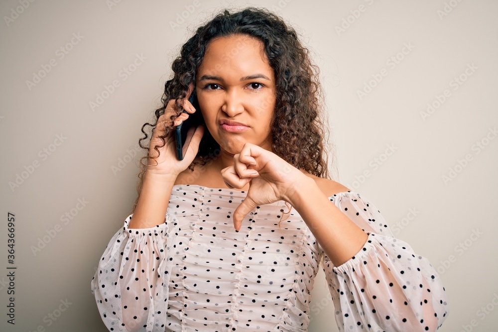 Young beautiful woman with curly hair having conversation talking on the smartphone with angry face, negative sign showing dislike with thumbs down, rejection concept Young beautiful woman with curly hair having conversation talking on the smartphone with angry face, negative sign showing dislike with thumbs down, rejection concept