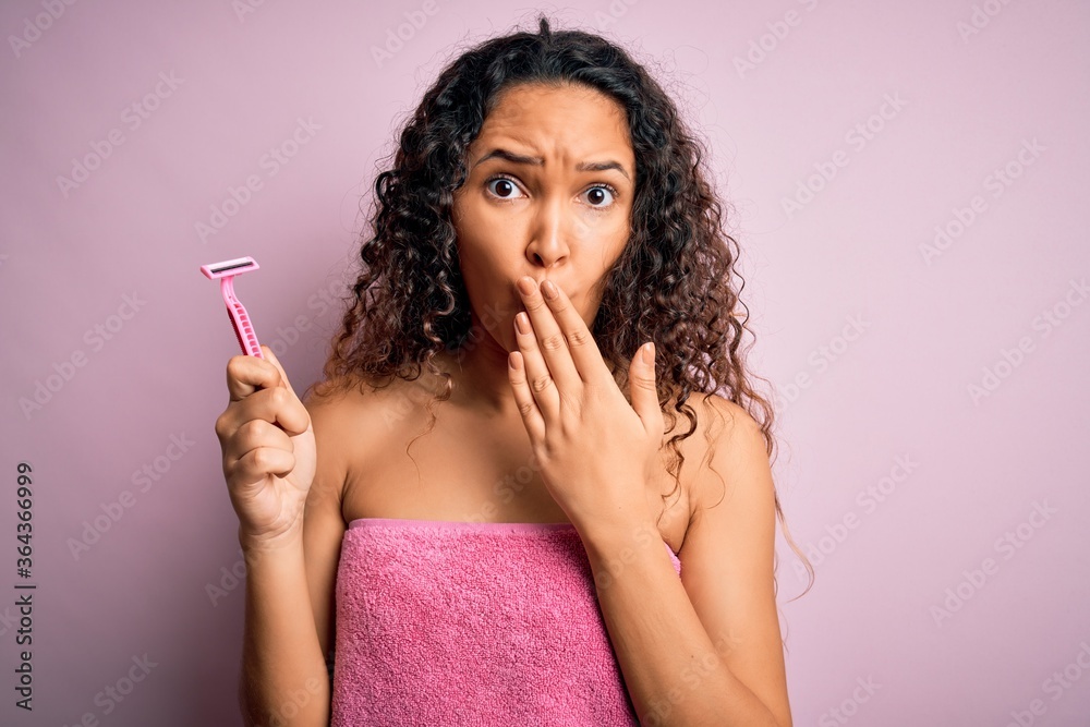 Young beautiful woman with curly hair wearing shower towel holding depilation razor cover mouth with hand shocked with shame for mistake, expression of fear, scared in silence, secret concept
