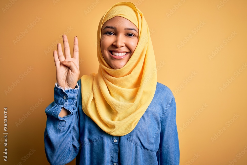 Young beautiful woman with curly hair wearing arab traditional hijab over yellow background showing and pointing up with fingers number four while smiling confident and happy. Young beautiful woman with curly hair wearing arab traditional hijab over yellow background showing and pointing up with fingers number four while smiling confident and happy.