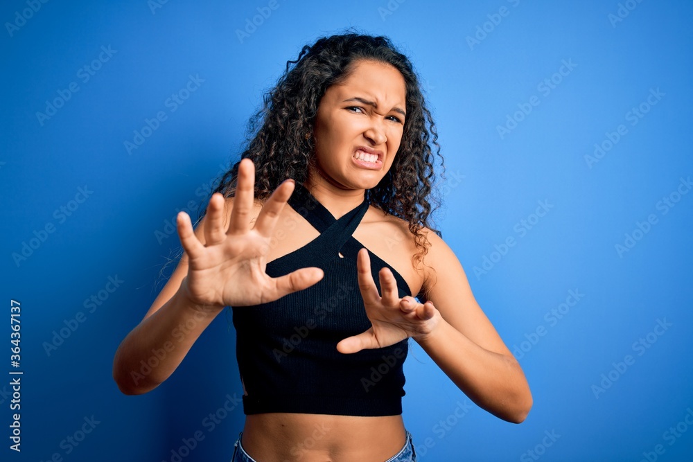 Young beautiful woman with curly hair wearing casual t-shirt standing over blue background disgusted expression, displeased and fearful doing disgust face because aversion reaction. With hands raised