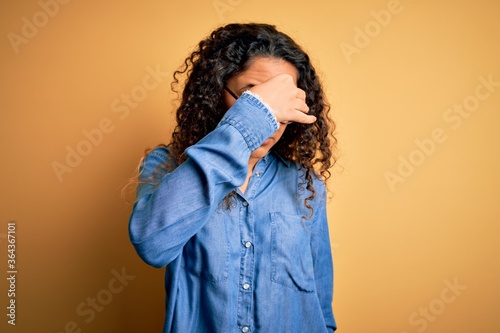 Beautiful woman with curly hair wearing casual denim shirt and glasses over yellow background tired rubbing nose and eyes feeling fatigue and headache. Stress and frustration concept.