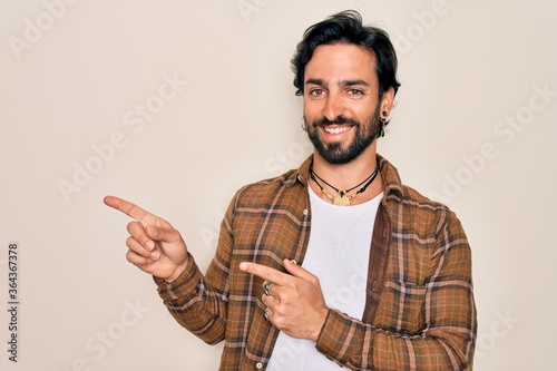Young handsome hispanic bohemian man wearing hippie style standing over isolated background smiling and looking at the camera pointing with two hands and fingers to the side.