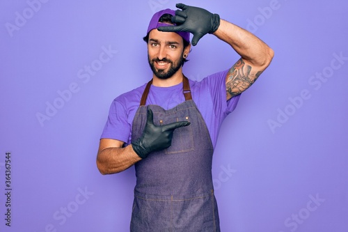 Young handsome hispanic man with bear wearing professional apron working as tattoo artist smiling making frame with hands and fingers with happy face. Creativity and photography concept. © Krakenimages.com
