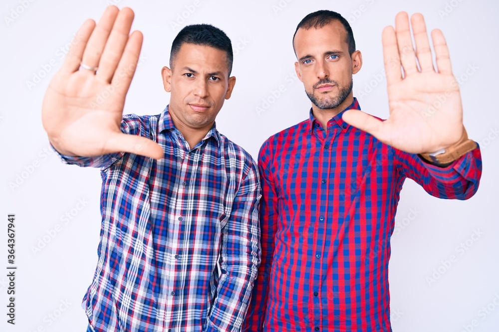 Young gay couple of two men wearing casual clothes doing stop sing with palm of the hand. warning expression with negative and serious gesture on the face. Young gay couple of two men wearing casual clothes doing stop sing with palm of the hand. warning expression with negative and serious gesture on the face.