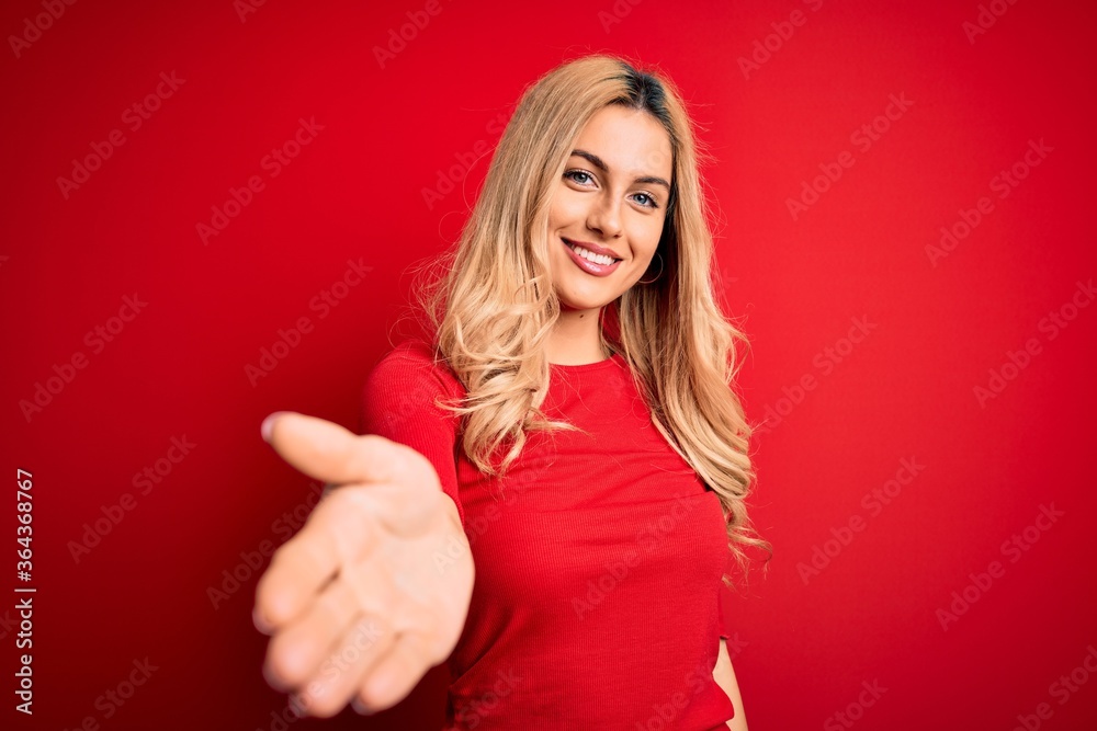 Young beautiful blonde woman wearing casual t-shirt standing over isolated red background smiling cheerful offering palm hand giving assistance and acceptance.