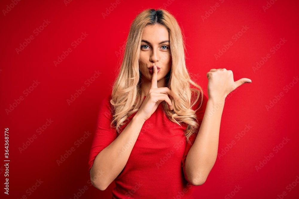 Young beautiful blonde woman wearing casual t-shirt standing over isolated red background asking to be quiet with finger on lips pointing with hand to the side. Silence and secret concept.