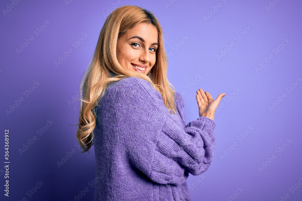 Young beautiful blonde woman wearing casual turtleneck sweater over purple background Inviting to enter smiling natural with open hand Young beautiful blonde woman wearing casual turtleneck sweater over purple background Inviting to enter smiling natural with open hand