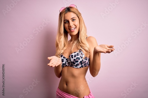Young beautiful blonde woman on vacation wearing bikini over isolated pink background smiling cheerful with open arms as friendly welcome, positive and confident greetings © Krakenimages.com