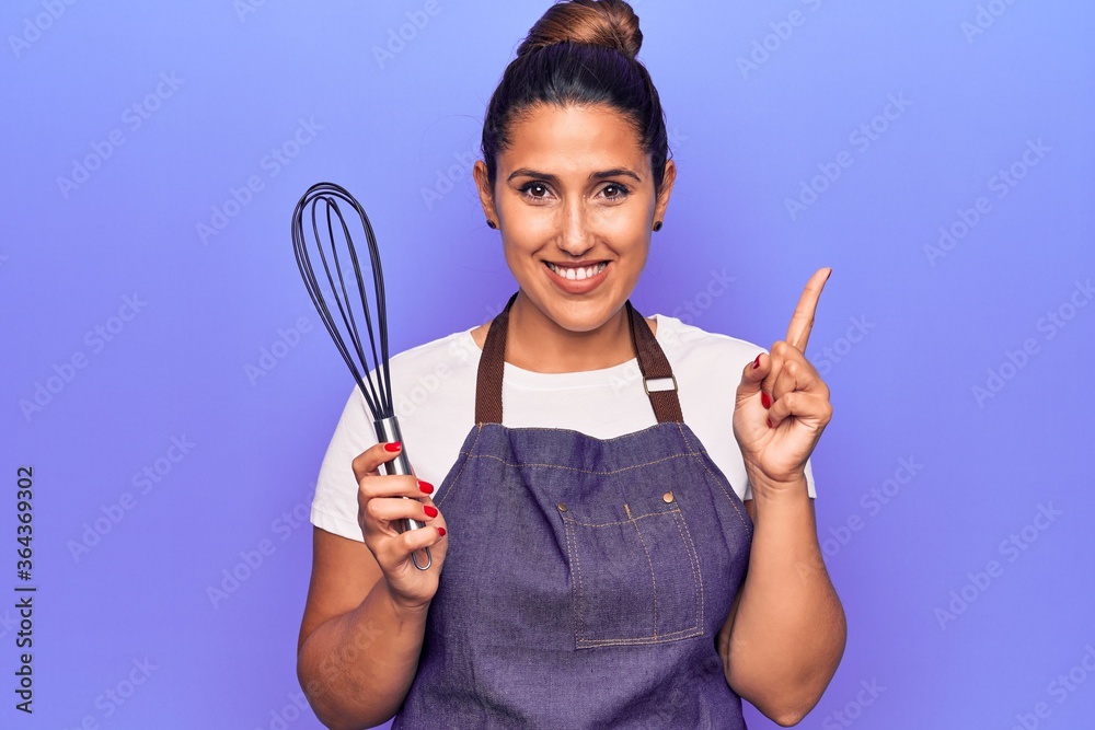 Young beautiful brunette woman wearing apron holding whisk smiling happy pointing with hand and finger to the side Young beautiful brunette woman wearing apron holding whisk smiling happy pointing with hand and finger to the side