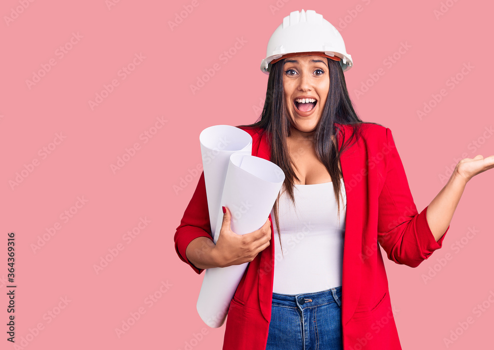 Young beautiful brunette woman wearing hardhat holding architect blueprint celebrating victory with happy smile and winner expression with raised hands Young beautiful brunette woman wearing hardhat holding architect blueprint celebrating victory with happy smile and winner expression with raised hands