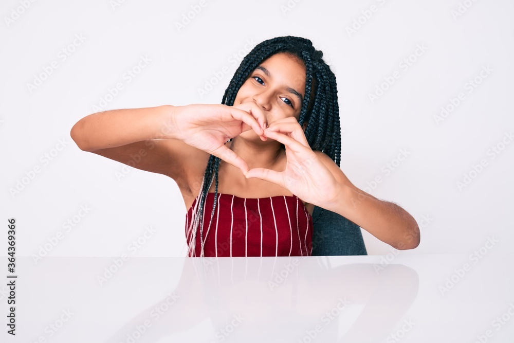 Young african american girl child with braids wearing casual clothes sitting on the table smiling in love doing heart symbol shape with hands. romantic concept.
