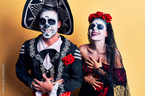 Young couple wearing mexican day of the dead costume over yellow smiling and laughing hard out loud because funny crazy joke with hands on body.