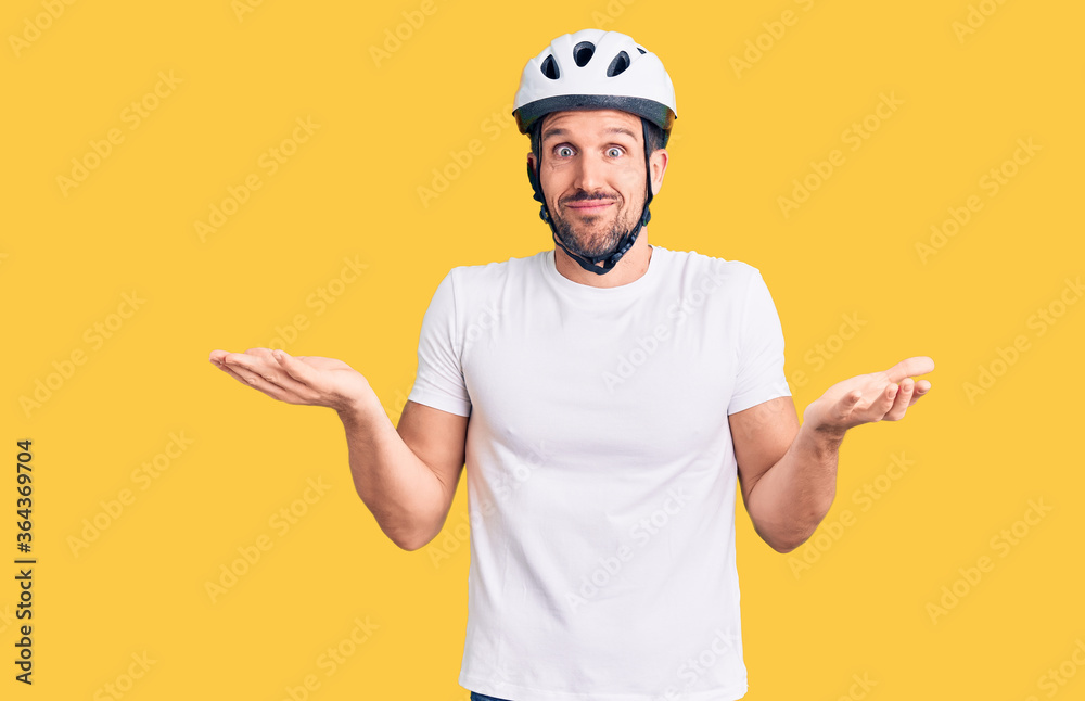 Young handsome man wearing bike helmet clueless and confused expression with arms and hands raised. doubt concept.