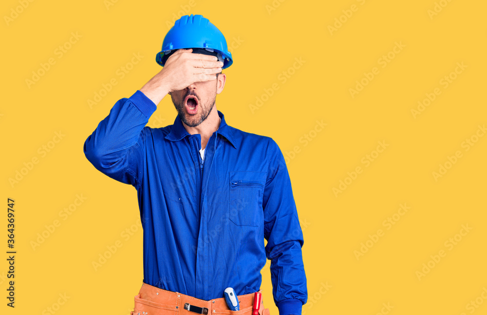 Young handsome man wearing worker uniform and hardhat smiling and laughing with hand on face covering eyes for surprise. blind concept.
