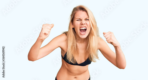 Billede på lærred Young beautiful blonde woman wearing bikini angry and mad raising fists frustrated and furious while shouting with anger