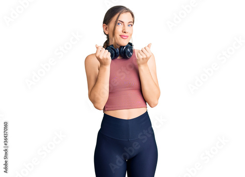 Beautiful caucasian young woman wearing gym clothes and using headphones doing money gesture with hands, asking for salary payment, millionaire business