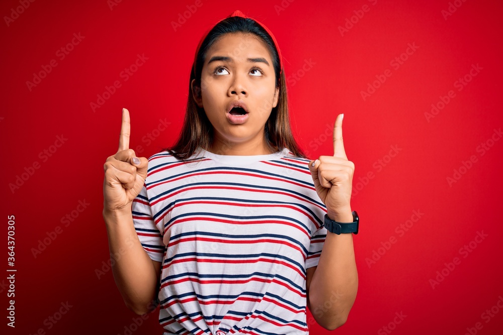 Young beautiful asian girl wearing casual striped t-shirt over isolated red background amazed and surprised looking up and pointing with fingers and raised arms. Young beautiful asian girl wearing casual striped t-shirt over isolated red background amazed and surprised looking up and pointing with fingers and raised arms.