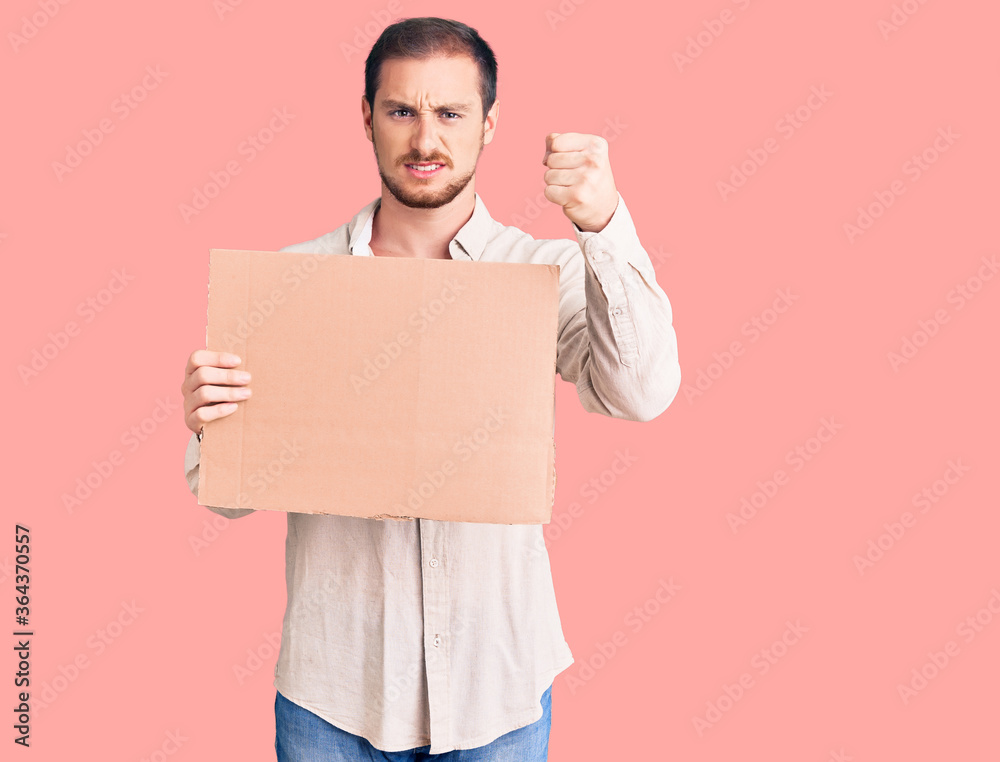 Young handsome caucasian man holding empty cardboard banner annoyed and frustrated shouting with anger, yelling crazy with anger and hand raised Young handsome caucasian man holding empty cardboard banner annoyed and frustrated shouting with anger, yelling crazy with anger and hand raised