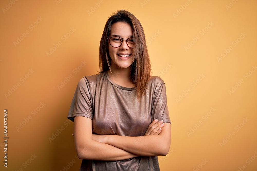 Obraz premium Beautiful brunette girl wearing casual t-shirt and glasses over isolated yellow background happy face smiling with crossed arms looking at the camera. Positive person.