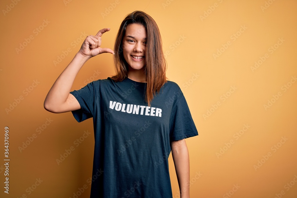 Young beautiful brunette girl doing volunteering wearing t-shirt with volunteer message word smiling and confident gesturing with hand doing small size sign with fingers looking and the camera. Young beautiful brunette girl doing volunteering wearing t-shirt with volunteer message word smiling and confident gesturing with hand doing small size sign with fingers looking and the camera.