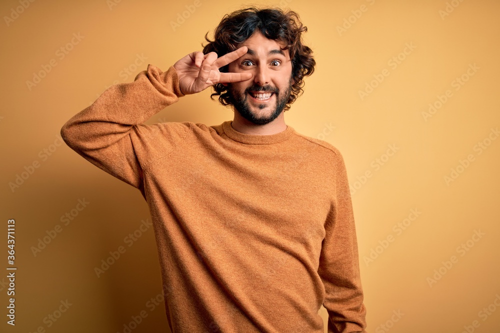 Young handsome man with beard wearing casual sweater standing over yellow background Doing peace symbol with fingers over face, smiling cheerful showing victory Young handsome man with beard wearing casual sweater standing over yellow background Doing peace symbol with fingers over face, smiling cheerful showing victory