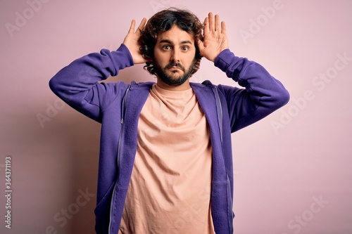 Young handsome sporty man with beard wearing casual sweatshirt over pink background Doing bunny ears gesture with hands palms looking cynical and skeptical. Easter rabbit concept. © Krakenimages.com