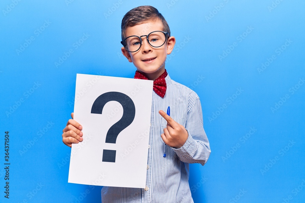 Cute blond kid wearing nerd bow tie and glasses holding question mark smiling happy pointing with hand and finger Cute blond kid wearing nerd bow tie and glasses holding question mark smiling happy pointing with hand and finger