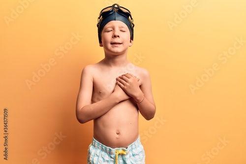 Cute blond kid wearing swimwear and swimmer glasses smiling with hands on chest with closed eyes and grateful gesture on face. health concept. © Krakenimages.com