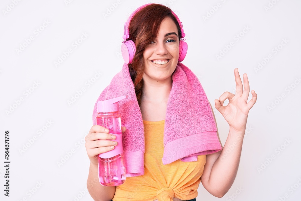 Young beautiful woman wearing towel and headphones holding bottle of water doing ok sign with fingers, smiling friendly gesturing excellent symbol Young beautiful woman wearing towel and headphones holding bottle of water doing ok sign with fingers, smiling friendly gesturing excellent symbol