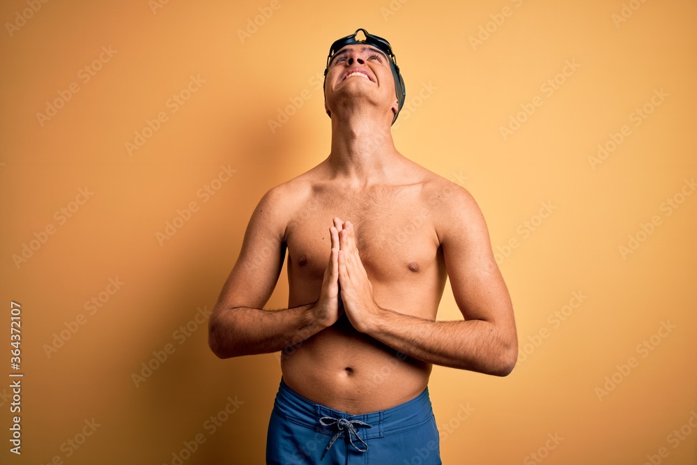 Young handsome man shirtless wearing swimsuit and swim cap over isolated yellow background begging and praying with hands together with hope expression on face very emotional and worried. Begging.