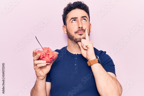 Young hispanic man holding ice cream serious face thinking about question with hand on chin, thoughtful about confusing idea