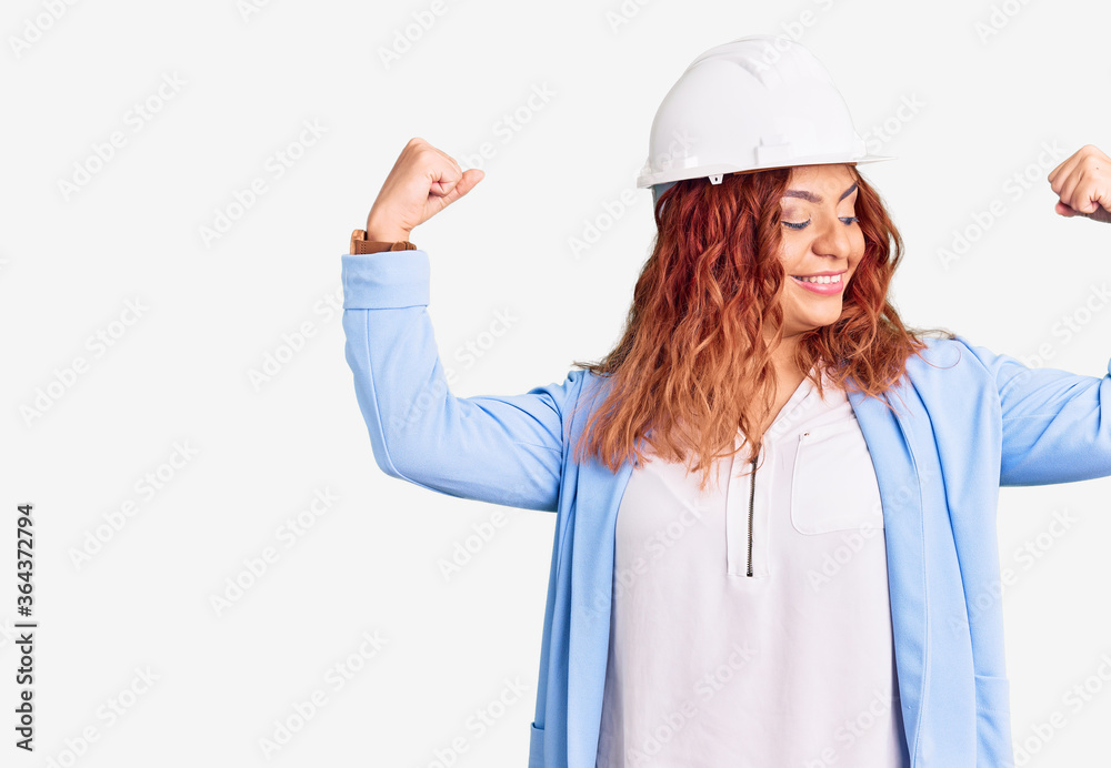 Young latin woman wearing architect hardhat showing arms muscles smiling proud. fitness concept. Young latin woman wearing architect hardhat showing arms muscles smiling proud. fitness concept.