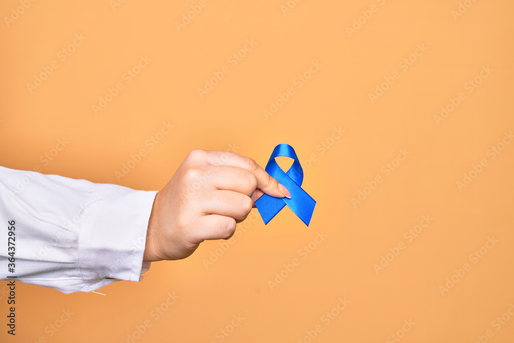 Hand of caucasian young woman holding blue cancer ribbon over isolated yellow background Hand of caucasian young woman holding blue cancer ribbon over isolated yellow background
