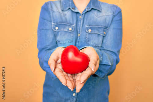 Woman holding red heart with two hands standing over isolated yellow background