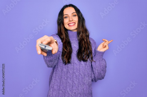 Obraz na plátně Young beautiful woman using television remote control over isolated purple backg