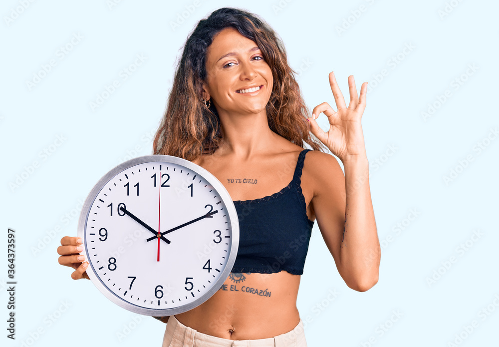 Young hispanic woman with tattoo holding big clock doing ok sign with fingers, smiling friendly gesturing excellent symbol Young hispanic woman with tattoo holding big clock doing ok sign with fingers, smiling friendly gesturing excellent symbol