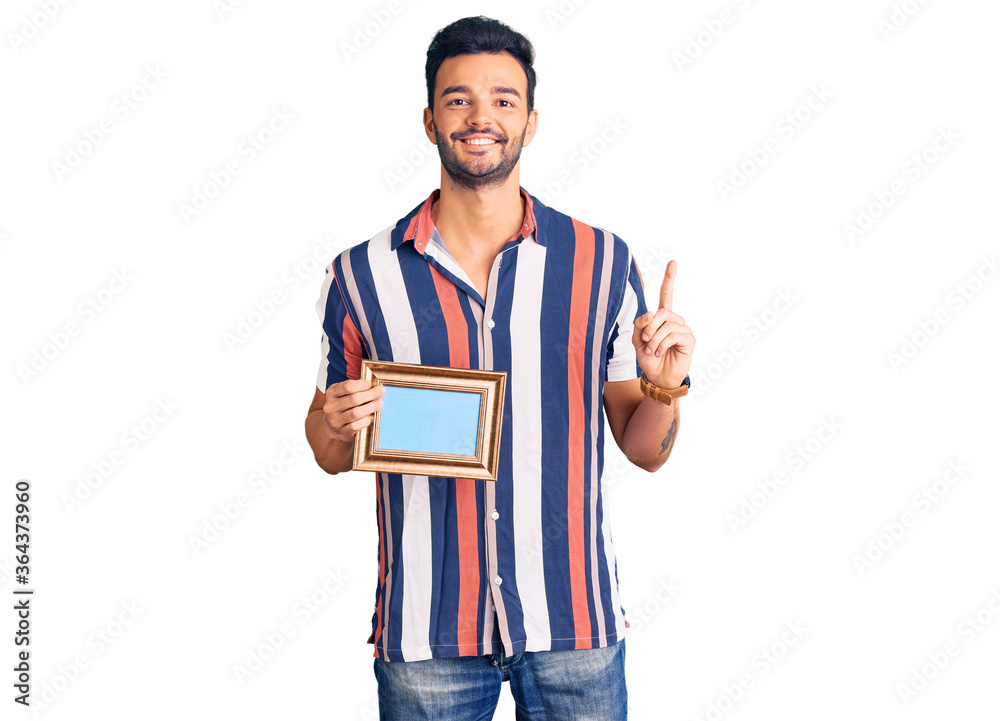 Young handsome hispanic man holding empty frame surprised with an idea or question pointing finger with happy face, number one Young handsome hispanic man holding empty frame surprised with an idea or question pointing finger with happy face, number one