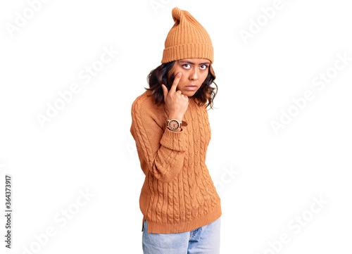 Young beautiful mixed race woman wearing wool sweater and winter hat pointing to the eye watching you gesture, suspicious expression photo