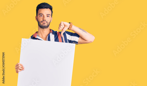 Young handsome hispanic man holding cardboard banner with blank space with angry face, negative sign showing dislike with thumbs down, rejection concept