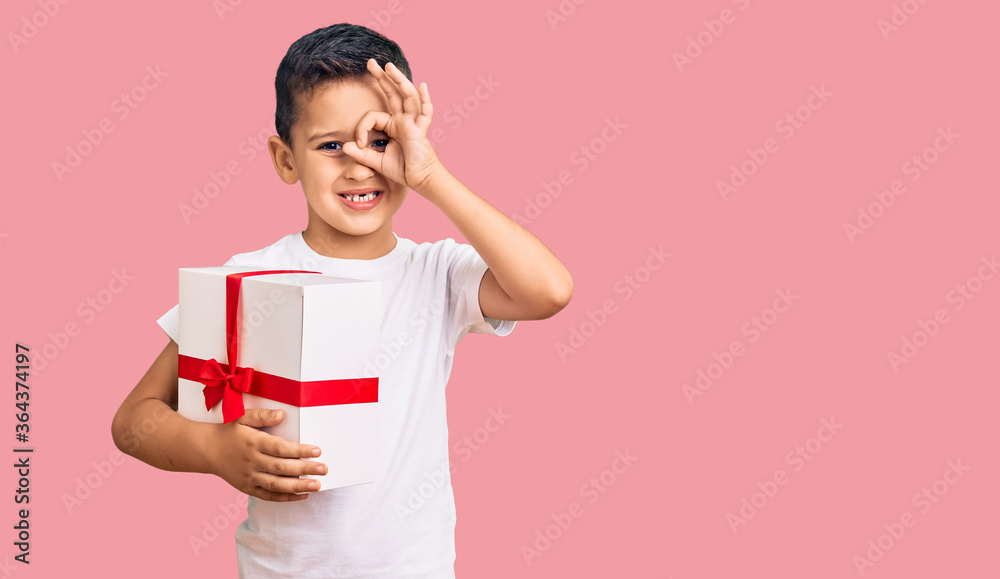 Little cute boy kid holding gift smiling happy doing ok sign with hand on eye looking through fingers