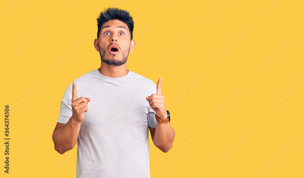 Handsome latin american young man wearing casual tshirt amazed and surprised looking up and pointing with fingers and raised arms. Handsome latin american young man wearing casual tshirt amazed and surprised looking up and pointing with fingers and raised arms.